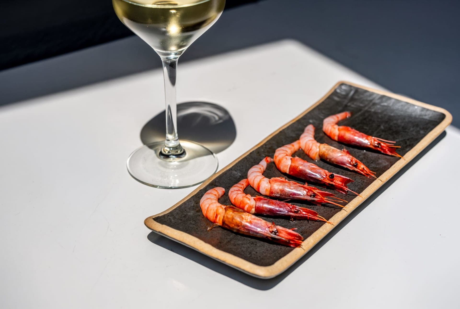 Gambes vermelles a la planxa en un plat fosc al costat d’una copa de vi blanc a La Subhasta.